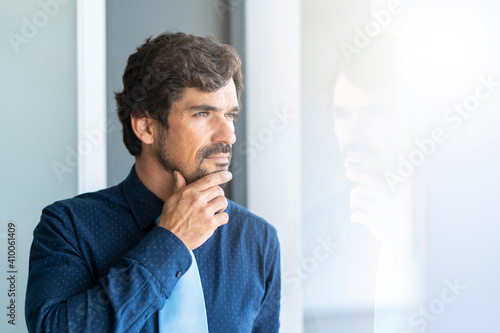 Man standing with hand on his chin looking through the window. Successful male portrait with a happy thinking expression face