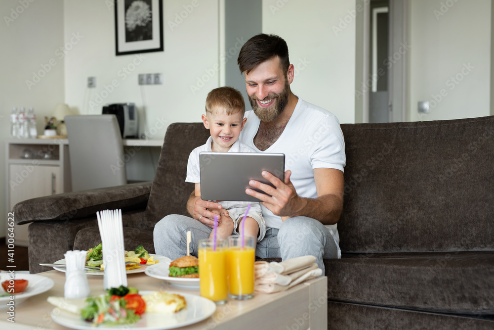 Father and son are having breakfast in a hotel room and looking at their laptop. Father and son are having breakfast in a hotel room and looking at their laptop.