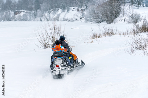 Father rides his son on a snowmobile along the bank of a frozen river