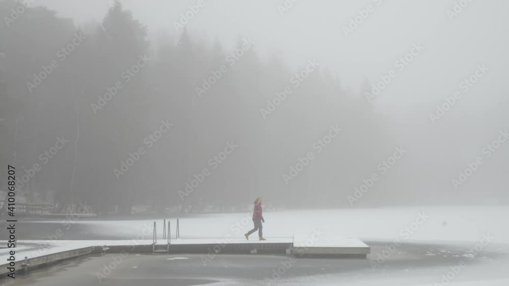 Tracking sideways shot of a Scandinavian woman walking along boardwalk over lake in wintery and moody national park in Finland with steam rising from the water. Tracking sideways shot of a Scandinavian woman walking along boardwalk over lake in wintery and moody national park in Finland with steam rising from the water.