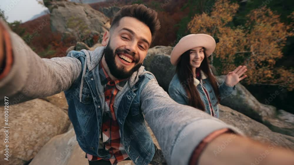 A young man is shooting a video on the front camera. A smiling woman is standing near his and waving her hand. Mountain landscape in the background. 4K. A young man is shooting a video on the front camera. A smiling woman is standing near his and waving her hand. Mountain landscape in the background. 4K.