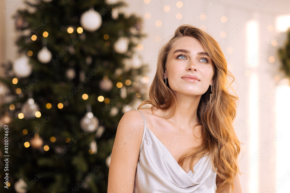 Portrait of a young pretty and dreamy blonde woman with a silver dress against the backdrop of the Christmas tree.