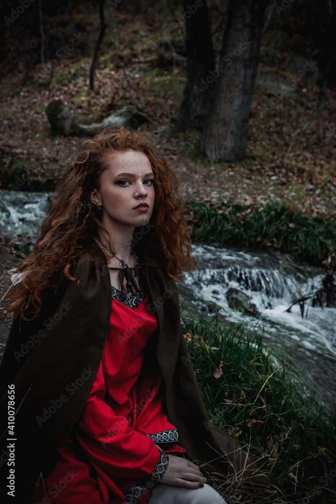 Fototapeta premium Red-haired woman in a red dress in a historical Celtic costume in the autumn forest