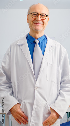 Portrait of elderly man surgeon standing in front of camera smiling in medical conference meeting office. Team of doctors working in background talking about symptoms of disease in clinic room