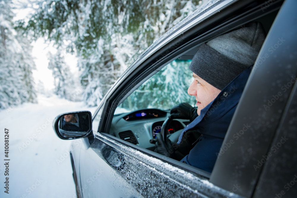 Man in warm winter clothes sitting in car. Snowy winter country road, car covered with ice, Beautiful forest under the snow. 