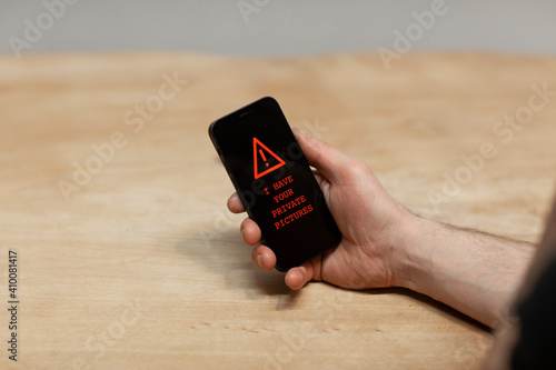 Red warning text saying ‘I have your private pictures’ on a hacked cell phone. The hand of he sextortion victim holding his phone while being desperate. Cybercrime, ransomware
