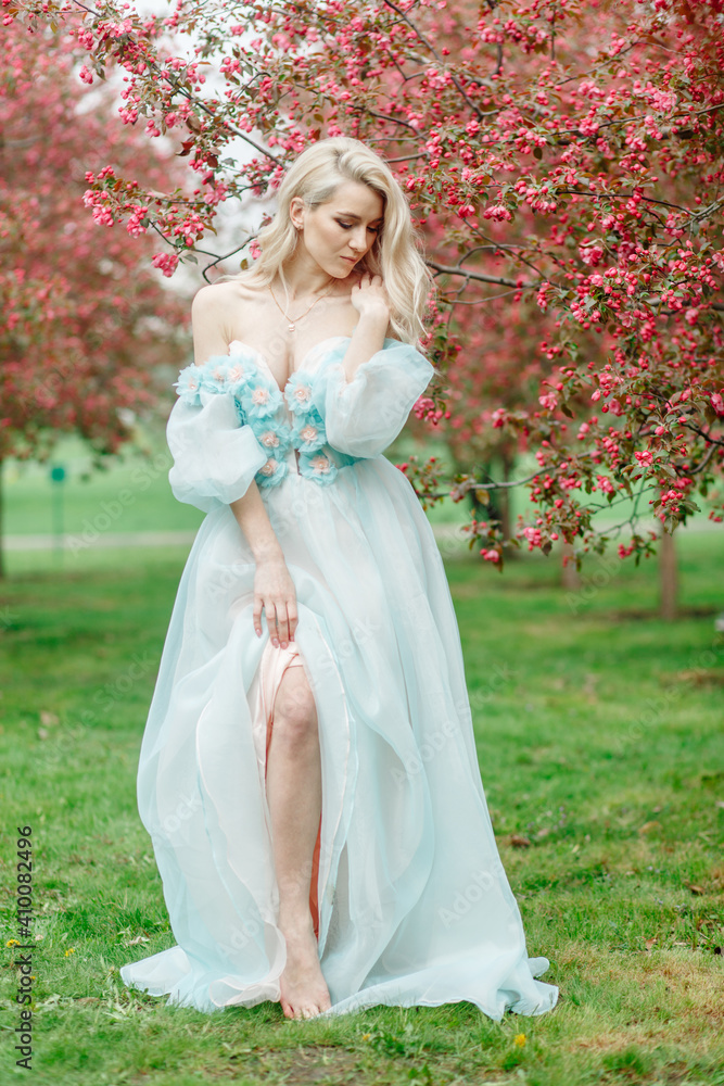 Blonde woman in spring in a pink blooming garden and in a beautiful blue dress Blonde woman in spring in a pink blooming garden and in a beautiful blue dress