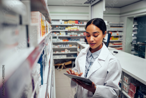 Young female pharmacist checking inventory of medicines in pharmacy using digital tablet wearing labcoat standing behind counter