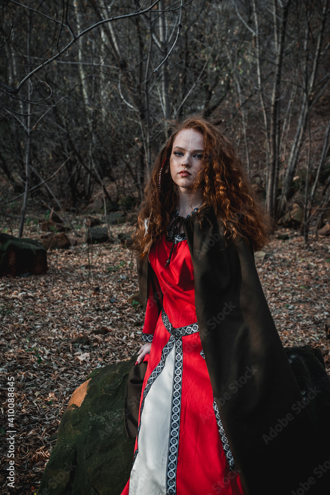 Red-haired woman in a red dress in a historical Celtic costume in the autumn forest