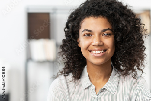 Headshot of a young elegant African American ethnic female with Afro curly hairstyle, beautiful smile and looking at the camera while standing against blurred home or office interior background