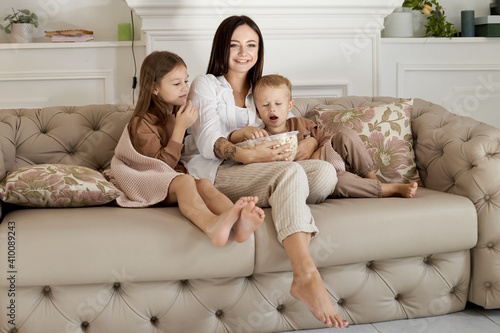 Mom sits on the couch with her son and daughter and watch a movie. A woman, a boy and a girl eat popcorn while watching a movie. The family is resting at home on the weekend