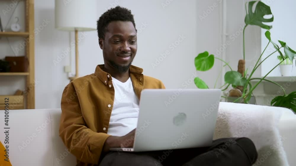 Man using laptop and smiling, sitting on couch at home room during pandemic spbas. Young American guy looks at computer screen and types with happy smile, sits on sofa in light interior. Modern person