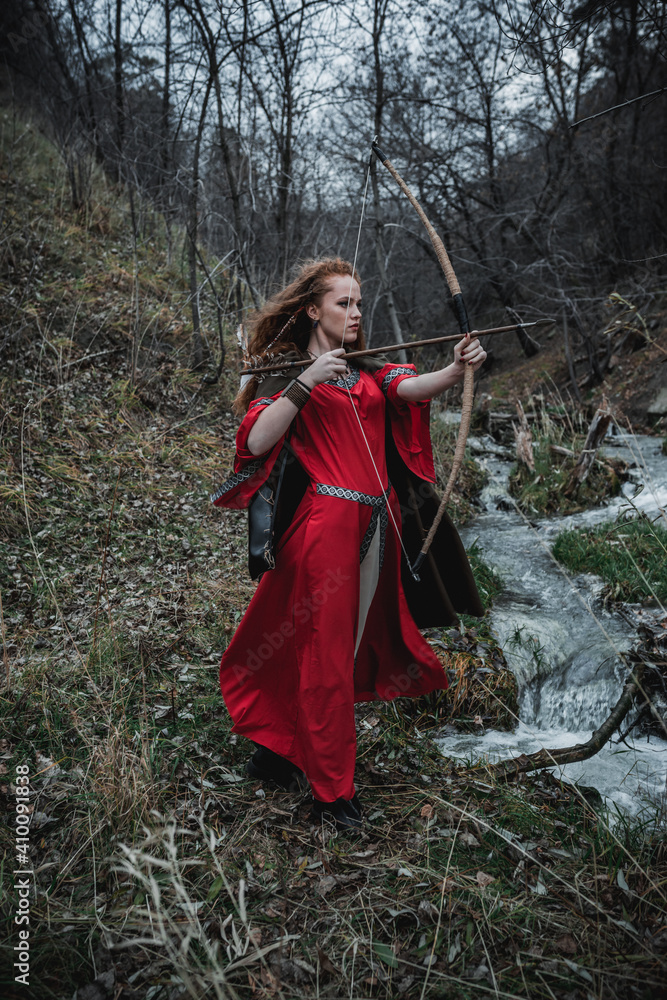 Red-haired woman in a red dress in a historical Celtic costume in the autumn forest Red-haired woman in a red dress in a historical Celtic costume in the autumn forest