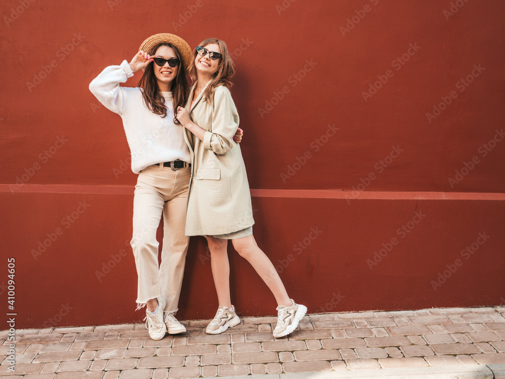 Two young beautiful smiling hipster female in trendy white sweater and coat. Sexy carefree women posing near red wall in the street in hat. Positive models having fun, going crazy in sunglasses Two young beautiful smiling hipster female in trendy white sweater and coat. Sexy carefree women posing near red wall in the street in hat. Positive models having fun, going crazy in sunglasses
