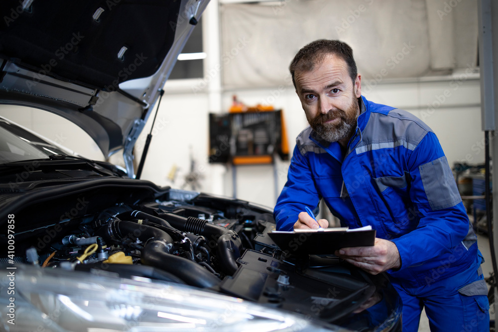 Professional middle aged caucasian car mechanic with checklist standing by vehicle engine area with hood open detecting malfunction. Professional middle aged caucasian car mechanic with checklist standing by vehicle engine area with hood open detecting malfunction.