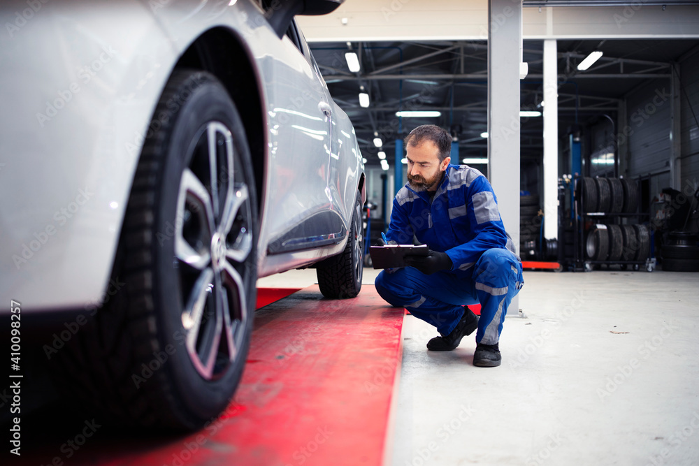 Professional middle aged bearded caucasian car mechanic doing visual inspection of vehicle in workshop and holding checklist. Automobile service and maintenance. Professional middle aged bearded caucasian car mechanic doing visual inspection of vehicle in workshop and holding checklist. Automobile service and maintenance.