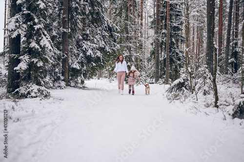 Happy family young mother and little cute girl in pink warm outwear walking having fun with red shiba inu dog in snowy white cold winter forest outdoors. Family sport vacation activities.