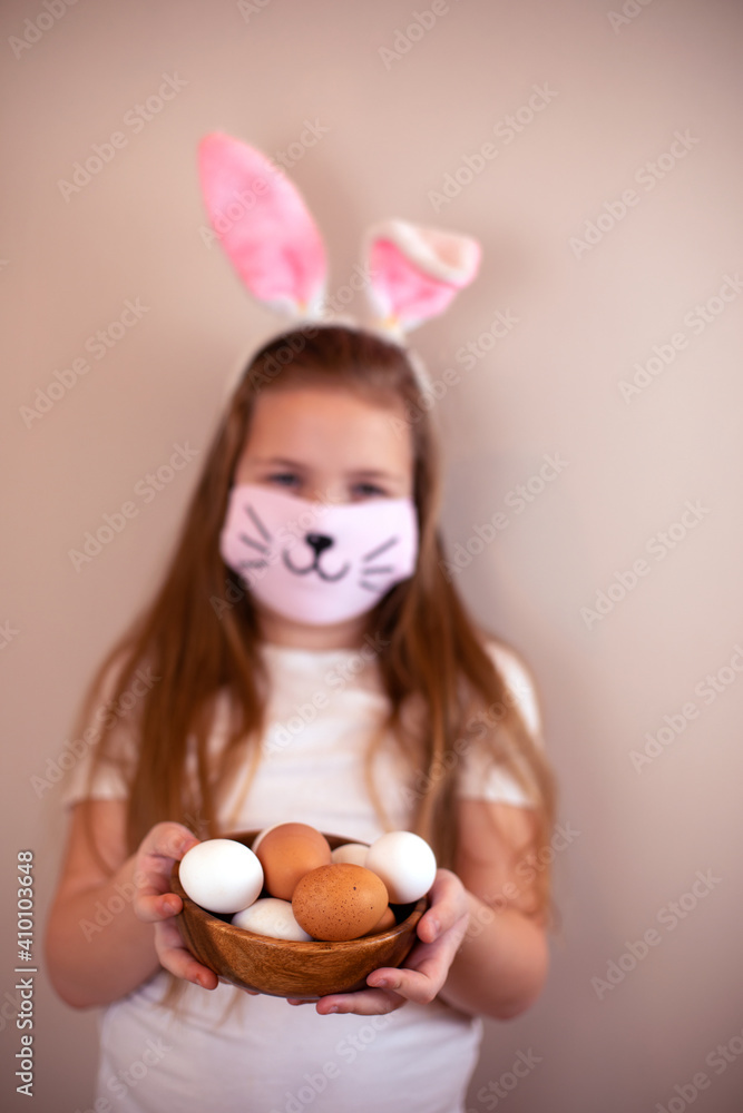 A little Caucasian girl with bunny ears and a medical mask with a painted nose and mustache on her face on a pink background with a bowl of white and brown eggs in her hands. Easter concept. A little Caucasian girl with bunny ears and a medical mask with a painted nose and mustache on her face on a pink background with a bowl of white and brown eggs in her hands. Easter concept.