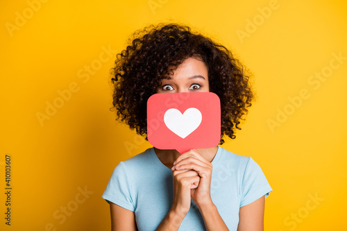 Photo of funky playful dark skin curly girl dressed blue t-shirt holding heart like card cover face isolated yellow color background photo