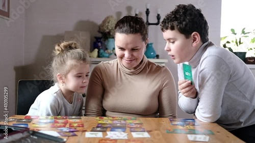 Happy mom is playing a board-based educational creative dice card game with the kids in the bright living room at the table.