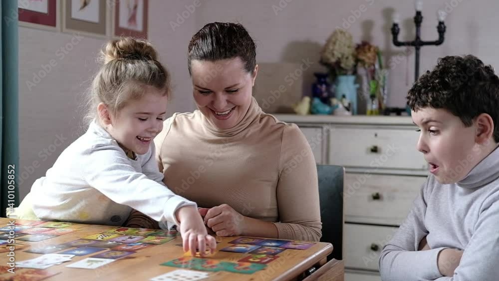 Happy kids play board-based educational creative dice card game with kids in bright living room at table. Mother helps children play Happy kids play board-based educational creative dice card game with kids in bright living room at table. Mother helps children play