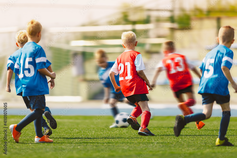 Obraz premium Young Footballers Kicking Football Match.Football Soccer Players Running with Ball. Soccer Players Running After the Ball. Kids in Soccer Red and Blue Uniforms. Soccer Stadium in the Background