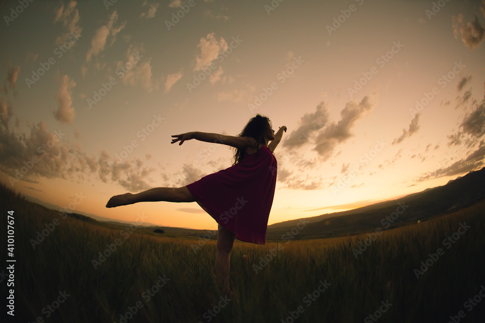 Young talented dancer girl raises her leg in the field of wild flowers during the gorgeous sunset