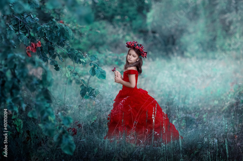 A child girl in a red dress in a wreath of berries on her head stands in a fairy forest