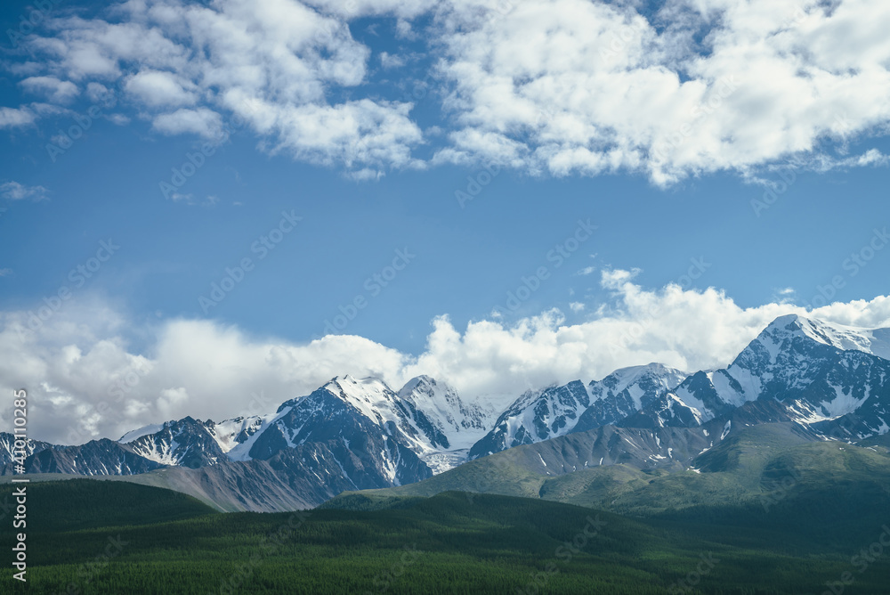 Awesome mountains landscape with sunlit high snowy pinnacle among low clouds in blue sky. Atmospheric highland scenery with snow-white big mountain top in sunlight. Wonderful snowy mountain range.