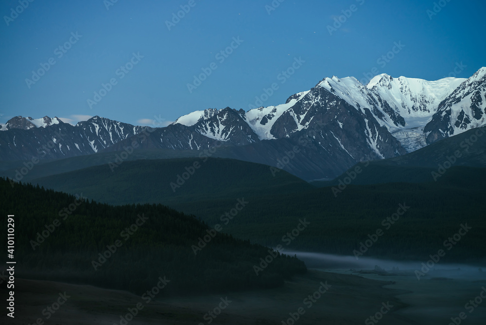 Atmospheric misty mountain landscape with great snow mountain top under twilight sky. Alpine misty scenery with big snowy mountains over forest in night. High snow pinnacle above forest hills in dusk.
