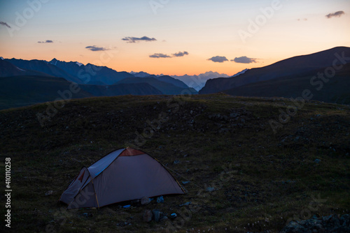 Colorful dawn landscape with tent on pass against lilac mountains under orange dawn sky with violet clouds. Tent on hill with view to great mountains silhouettes in sunset. Beautiful alpine sunrise.