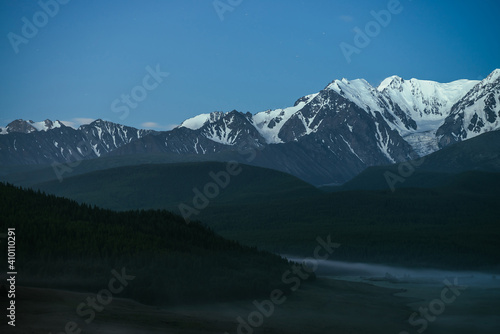 Atmospheric misty mountain landscape with great snow mountain top under twilight sky. Alpine misty scenery with big snowy mountains over forest in night. High snow pinnacle above forest hills in dusk.