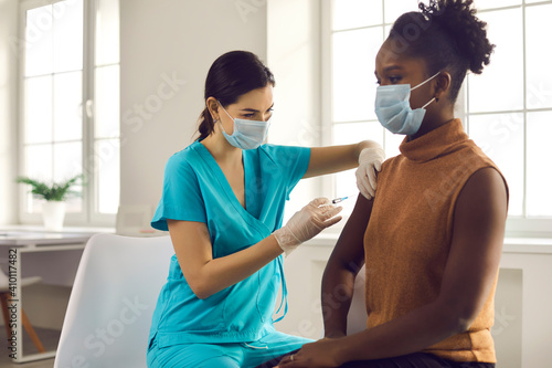 Young African American lady sitting at doctor's office and getting modern Covid 19 shot. Nurse in medical face mask and gloves holding syringe and giving female patient flu vaccine injection in arm