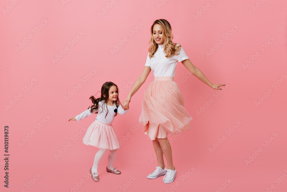 Positive blonde mom and dark-haired daughter in lush romantic skirts are dancing boogie woogie on pink background