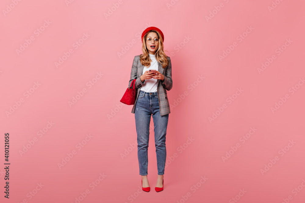 Amazed girl in white top, jeans and gray jacket is looking at camera. Blonde in glasses and hat holds smartphone and bag Amazed girl in white top, jeans and gray jacket is looking at camera. Blonde in glasses and hat holds smartphone and bag