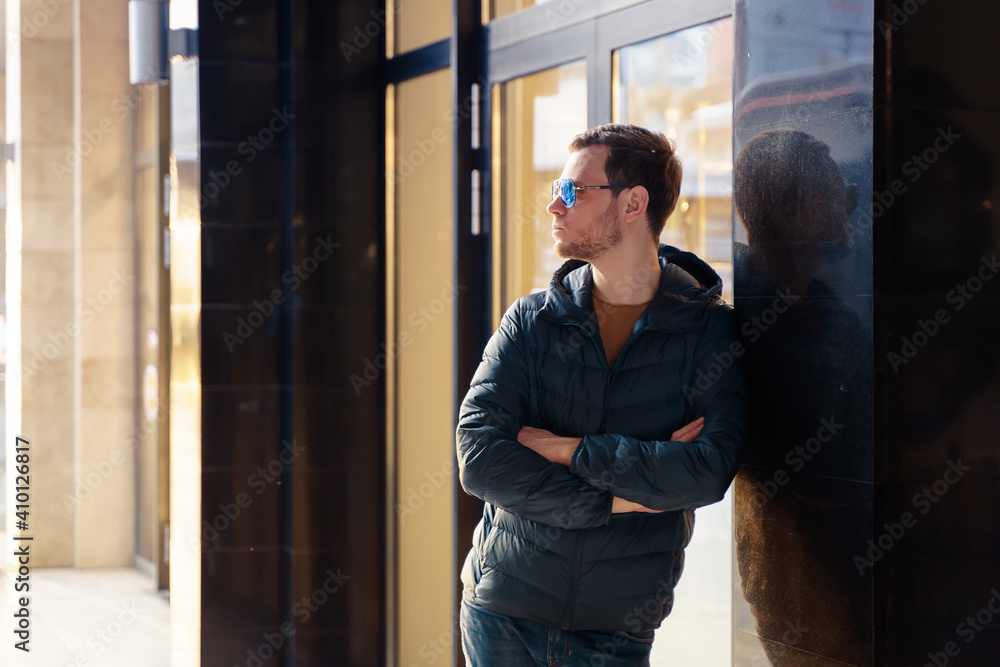 Young european man looking forward confidently through blue sunglasses standing with his arms crossed by modern glass wall in a mall or shopping centre. Backlit with golden sunset sun. Young european man looking forward confidently through blue sunglasses standing with his arms crossed by modern glass wall in a mall or shopping centre. Backlit with golden sunset sun.