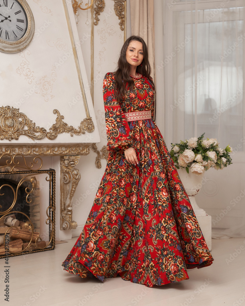 Elegant brunette young woman with long wavy hair in long colorful red dress standing and posing in bright luxury interior