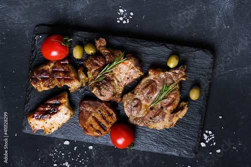  Variety of  grilled meat steaks steaks with spices, olives and tomatoes on black background. Top view