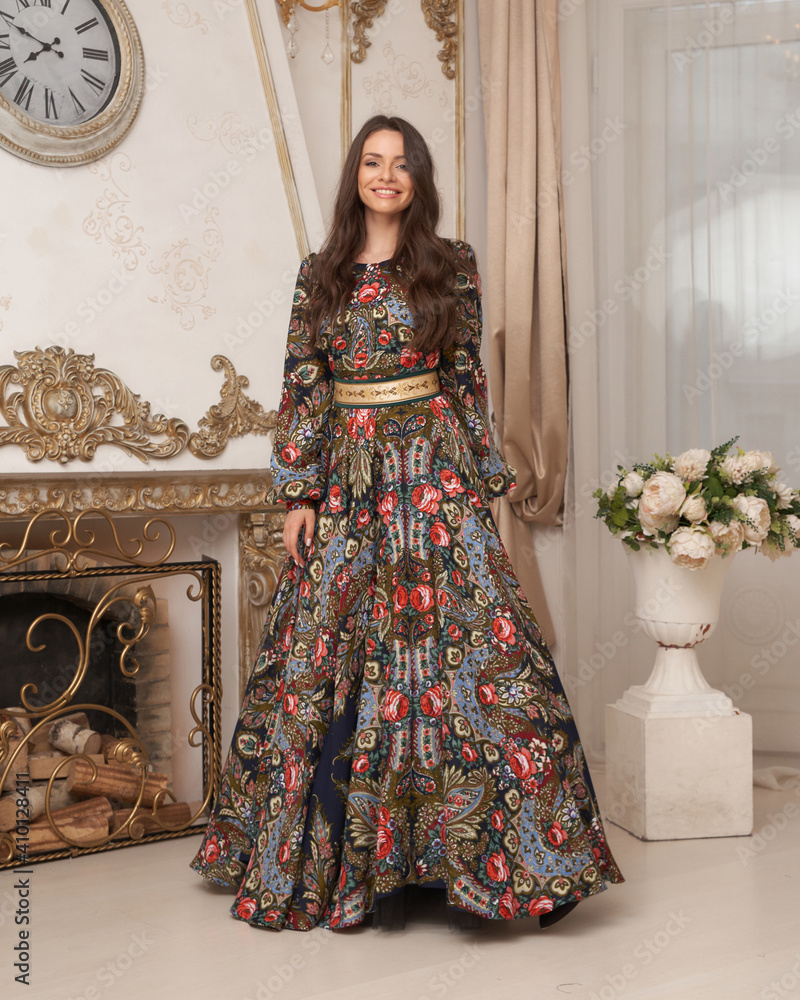 Elegant brunette young woman with long wavy hair in long colorful dress standing and posing in bright luxury interior
