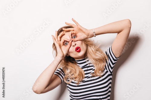 Funny girl with red lipstick fooling around and posing on white background. Woman folds her fingers in form of glasses and whistles