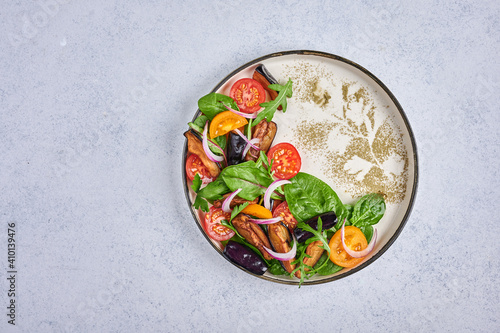 Homemade styling salad with fried eggplants, tomatoes, arugula, spinach, lettuce and sauce on a ceramic plate on light background, top view, copy space