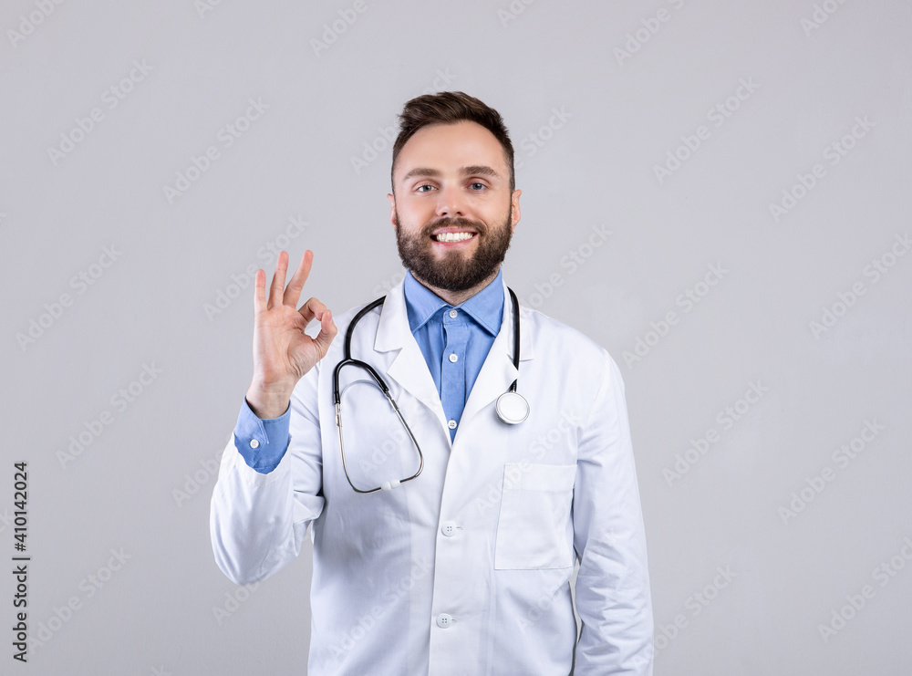 Portrait of happy young doctor in uniform showing okay gesture on grey studio background. Healthcare concept Portrait of happy young doctor in uniform showing okay gesture on grey studio background. Healthcare concept