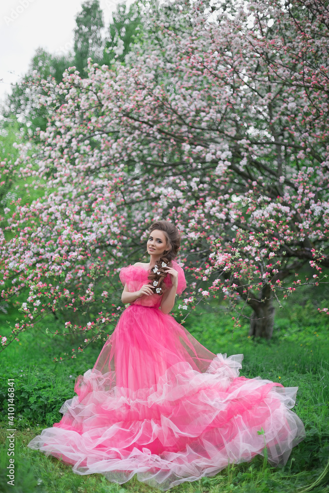 A beautiful young girl with long hair in a light pink ball gown walks through a blooming apple orchard