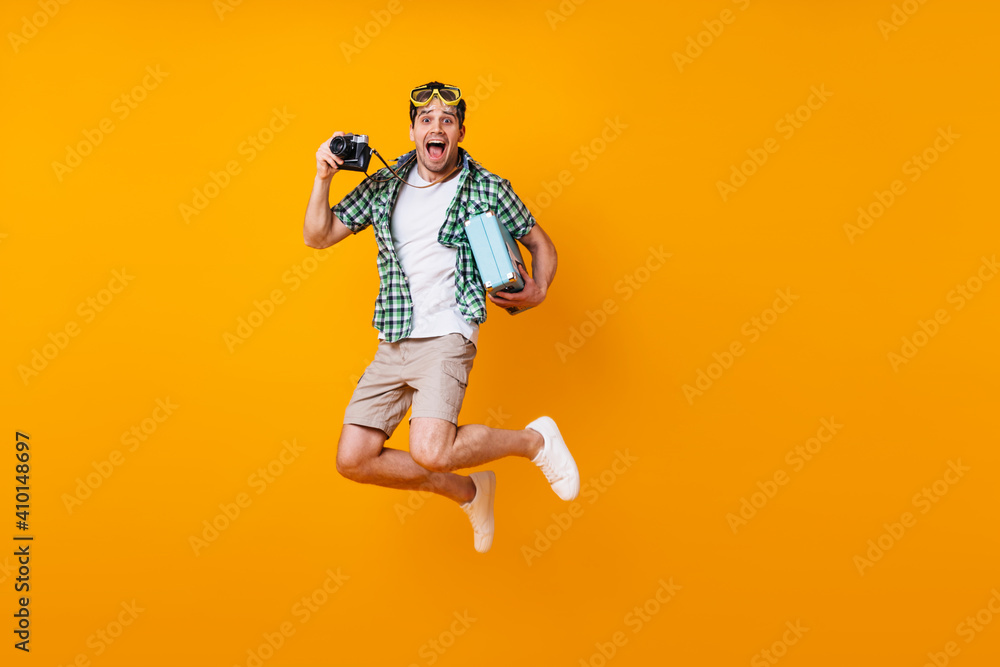 Emotional man in beige shorts, green shirt and white t-shirt jumping on orange background. Portrait of active guy holding retro camera and suitcase