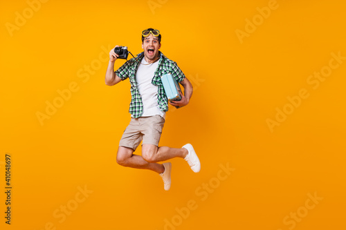 Emotional man in beige shorts, green shirt and white t-shirt jumping on orange background. Portrait of active guy holding retro camera and suitcase