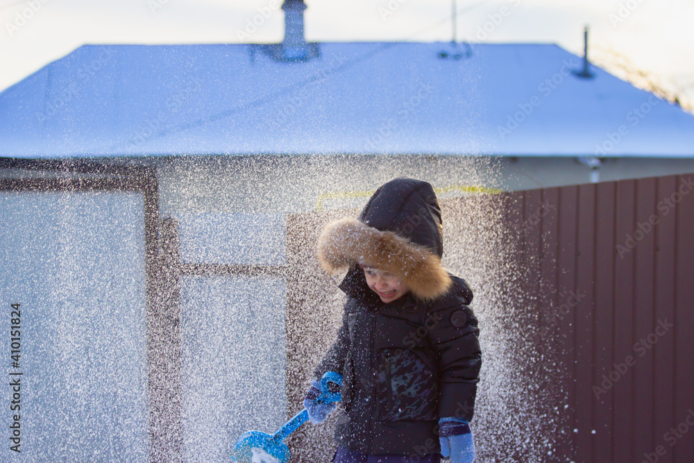 
little boy playing with snow and blue shovel in the yard of a private house. sunset in winter