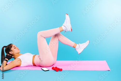 Obraz na płótnie Caucasian girl, during exercises on a yoga mat, raises her legs up and does exercise with a bicycle, shaking the press on a blue background. w salonie