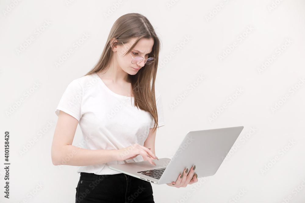 Naklejka premium Portrait of a beautiful young girl with blond hair in glasses and a white T-shirt working or studying on a laptop isolated on a white background. Concept of business woman. Casual blogger woman