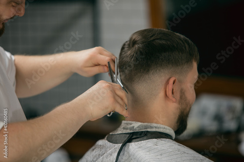 Close up master barber, stylist does the hairstyle to guy, young man. Professional occupation, male beauty concept. Cares of hair, mustache, beard of client. Soft colors and focus, vintage.