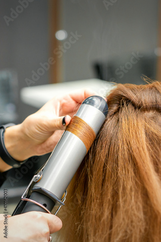 Professional hairdresser makes curls with a curling iron for a young woman with long red hair in a beauty salon